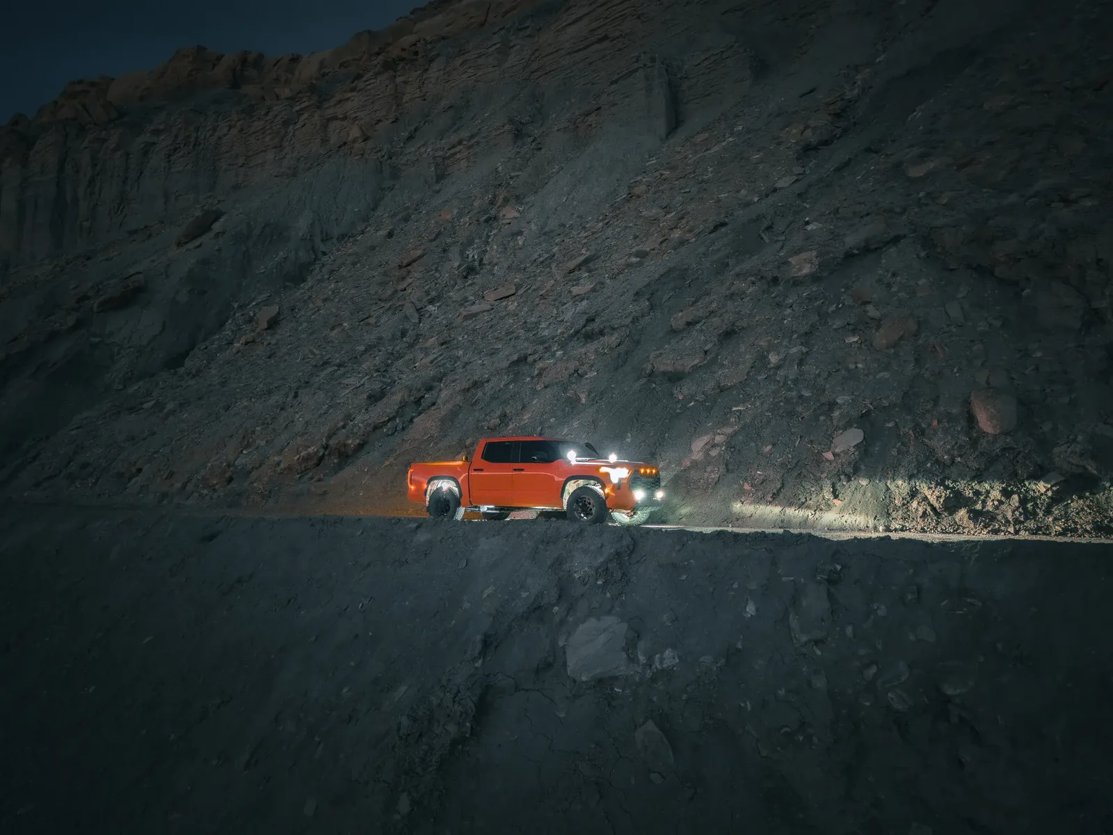 Orange Tundra at night, side profile on a dirt road through canyon walls, headlights lit.